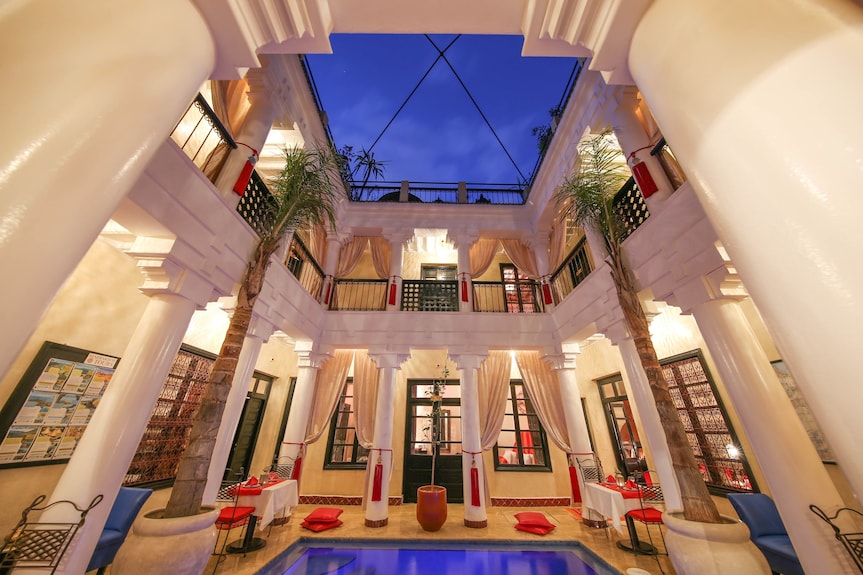 Riad Africa Marrakech courtyard with beige columns, blue pool, red cushions, tall windows, and evening sky