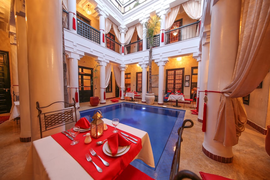 Riad Africa Marrakech indoor courtyard with blue tiled pool, yellow ochre walls, white columns, and red table settings