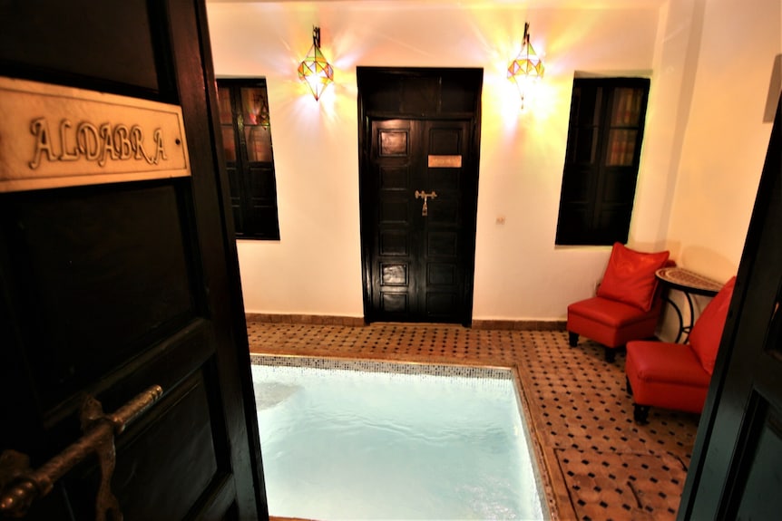 Indoor pool with mosaic tiled floor and walls, dark wooden doors and windows, lit Moroccan lanterns, and red cushioned chairs at Riad Africa Marrakech