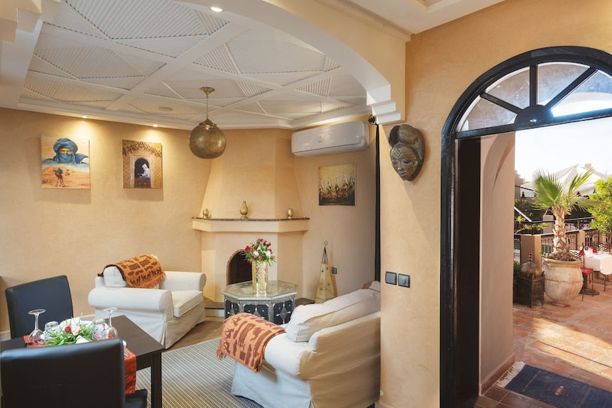 Riad Africa Marrakech living area with beige walls, white armchairs, patterned throws, decorative ceiling, and arched doorway leading to terrace