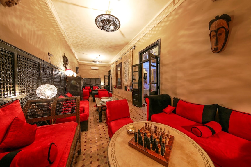 Riad Africa Marrakech lounge area with red and black upholstered sofas, wooden chess table, intricate tile flooring, and Moroccan wall decor