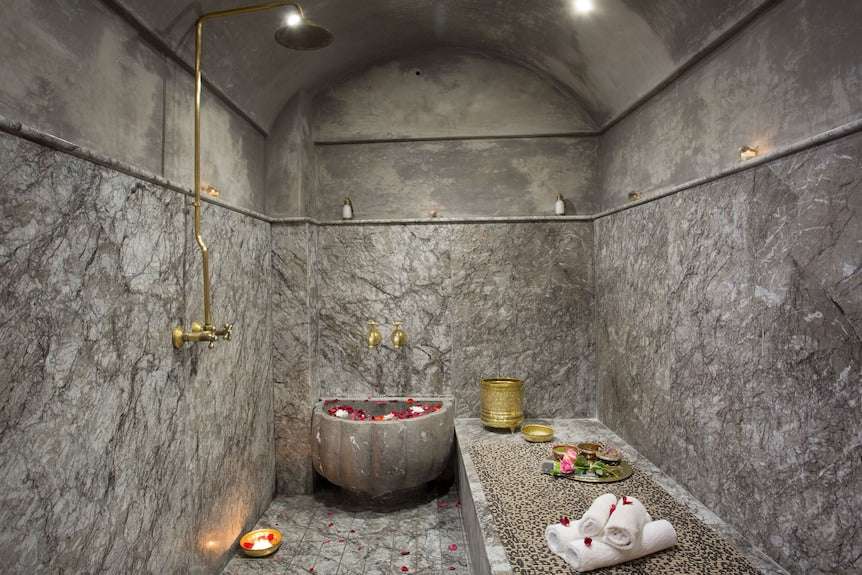 Riad Africa Marrakech traditional Moroccan hammam with grey marble walls, brass fixtures, stone basin with rose petals, and leopard print massage table