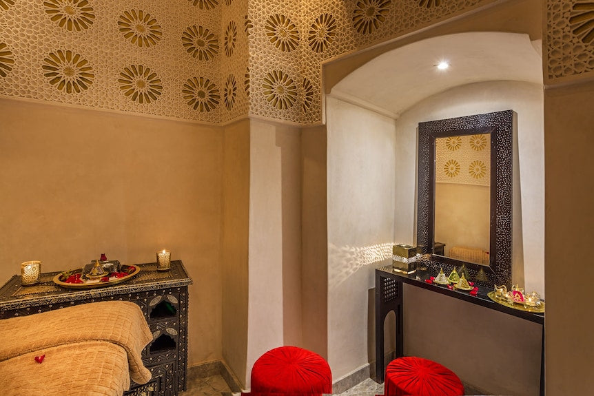Luxurious traditional Moroccan spa room with ornate carved ceiling, textured walls, red velvet stools, and dark wood furniture at Riad Africa Marrakech