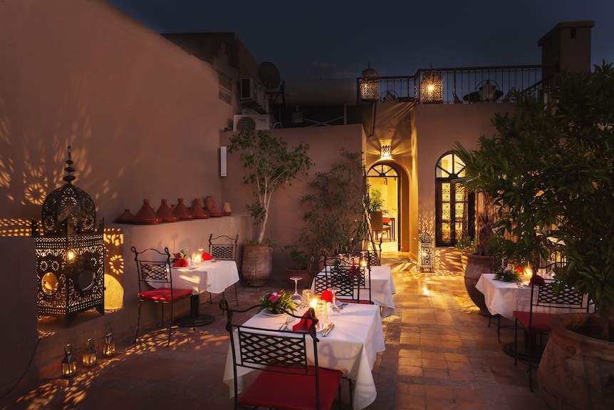 Outdoor dining area at Riad Africa Marrakech with iron chairs, white tablecloths, red cushions, Moroccan lanterns, and potted plants at night