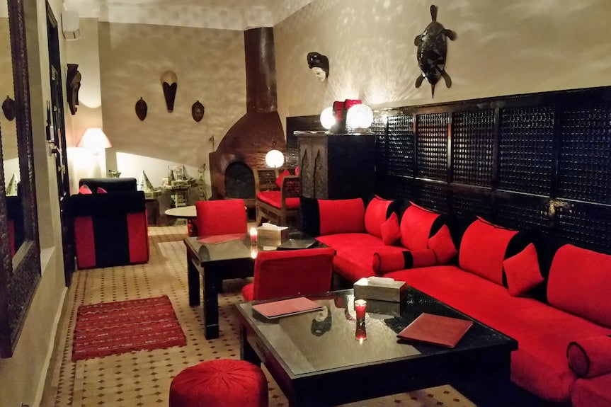 Riad Africa Marrakech lounge with red cushioned seating, black wood paneling, Moroccan decorative masks, and a traditional fireplace