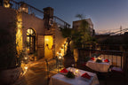 Riad Africa Marrakech outdoor terrace at dusk with warmly lit entrance, potted plants, iron railings, and two dining tables with red napkins