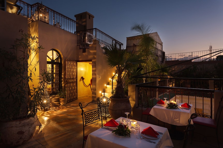 Riad Africa Marrakech outdoor terrace at dusk with warmly lit entrance, potted plants, iron railings, and two dining tables with red napkins