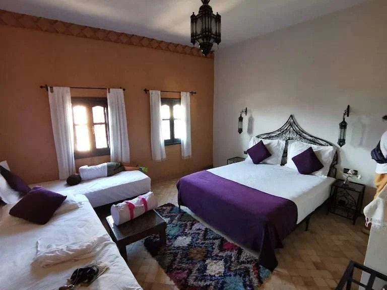 Bedroom at Riad Bouchedor in Ouarzazate featuring white linens with purple pillows and bed runner, terracotta walls, wooden windows, and traditional Moroccan rug