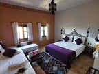 Bedroom at Riad Bouchedor in Ouarzazate featuring white linens with purple pillows and bed runner, terracotta walls, wooden windows, and traditional Moroccan rug