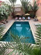 Outdoor swimming pool at Riad Bouchedor in Ouarzazate with surrounding palm trees, sun loungers, umbrellas, and terracotta walls