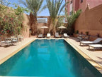 Riad Bouchedor in Ouarzazate outdoor swimming pool with turquoise water, surrounded by beige walls, palm trees, sun loungers and umbrellas