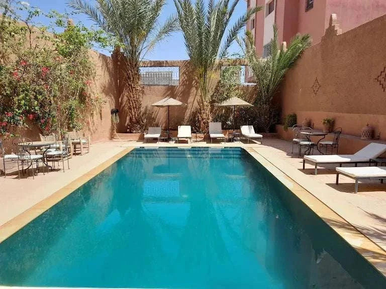 Riad Bouchedor in Ouarzazate outdoor swimming pool with turquoise water, surrounded by beige walls, palm trees, sun loungers and umbrellas