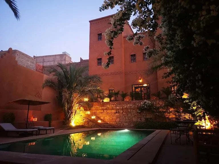 Riad Bouchedor in Ouarzazate featuring warm terracotta walls, illuminated pool, stone steps, and lush palm trees at dusk