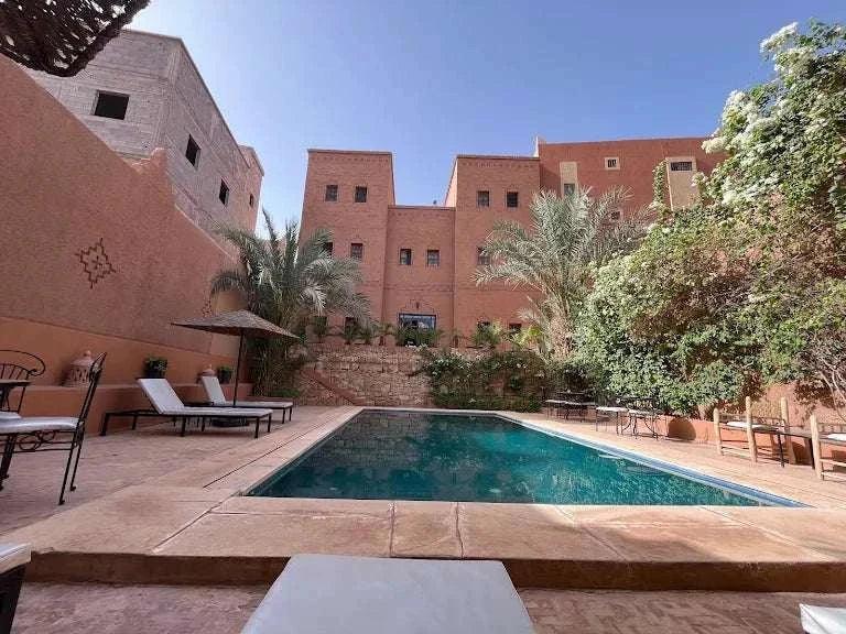 Riad Bouchedor in Ouarzazate featuring traditional Moroccan terracotta buildings surrounding a rectangular outdoor swimming pool with lounge chairs and greenery