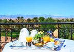 Riad Chay Boutique breakfast table with orange juice, white coffee pots, traditional teapot, bread, and flowers on a balcony with palm trees and mountains view