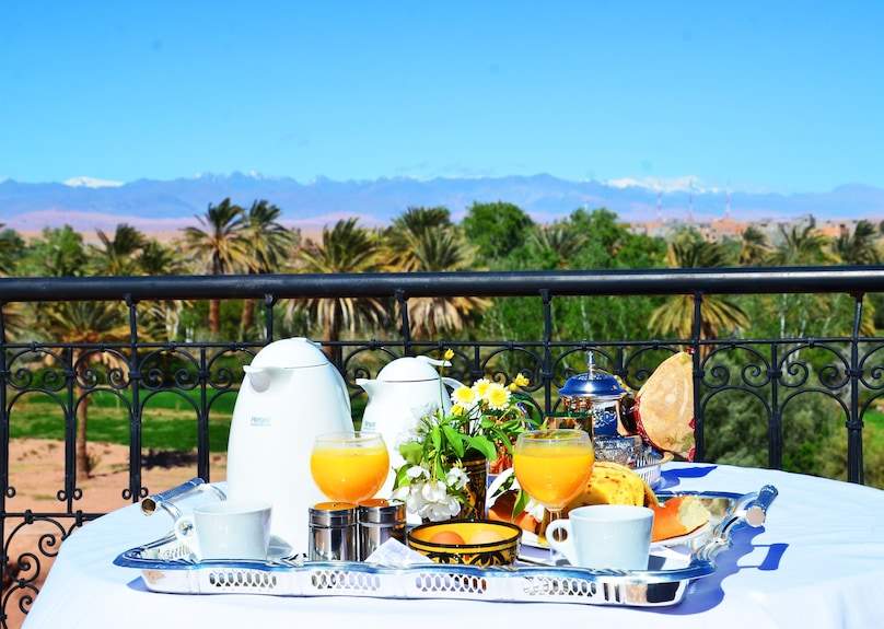 Riad Chay Boutique breakfast table with orange juice, white coffee pots, traditional teapot, bread, and flowers on a balcony with palm trees and mountains view