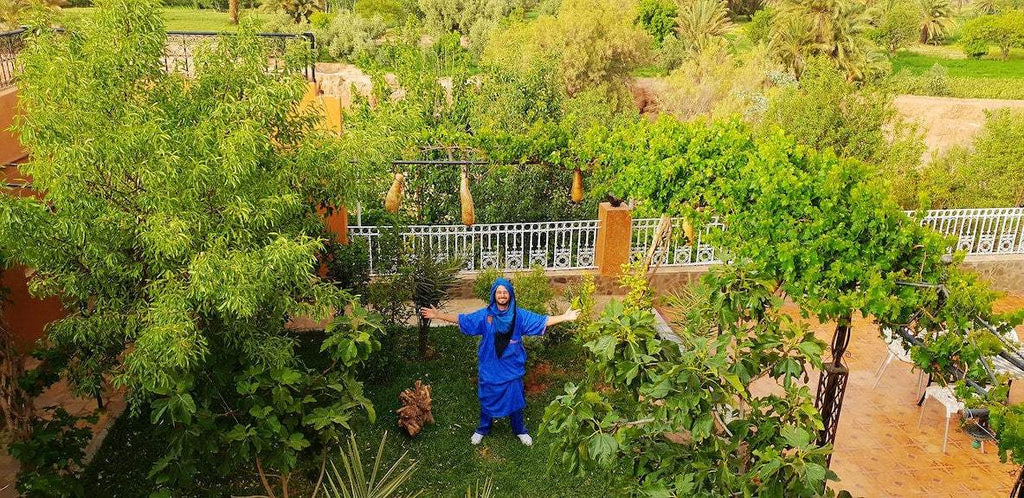 Riad Chay Boutique garden view with lush green trees, a person in blue traditional attire standing with arms open, and scenic Moroccan landscape