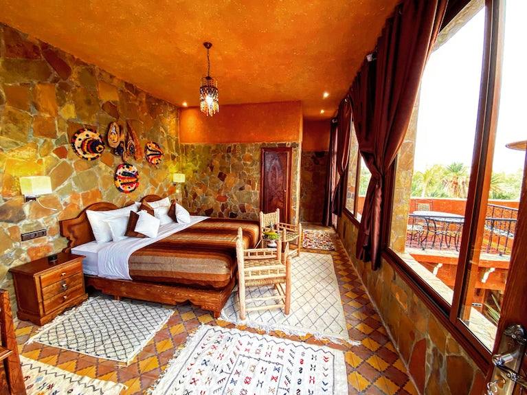 Riad Chay Boutique Moroccan-style bedroom with stone walls, warm tones, wooden furniture, patterned rugs, and large windows