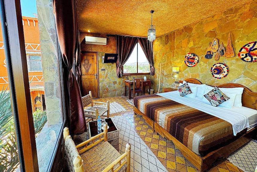 Riad Chay Boutique Moroccan-style bedroom with stone walls, wooden bed with striped brown bedding, woven chairs, and decorative wall hangings