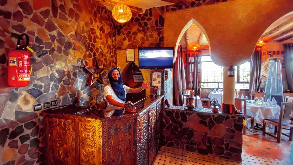 Reception area at Riad Chay Boutique featuring stone walls, carved wooden counter, warm lighting, and a smiling staff member