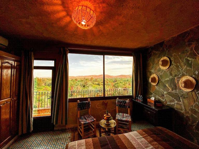 Riad Chay Boutique room with earth-toned decor, patterned tiled floor, wooden furniture, and large window overlooking lush greenery and distant hills