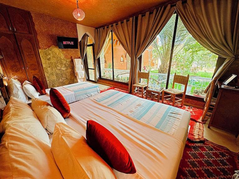 Riad Chay Boutique room with warm terracotta walls, two beds with striped blankets, red cushions, large windows, and wooden chairs