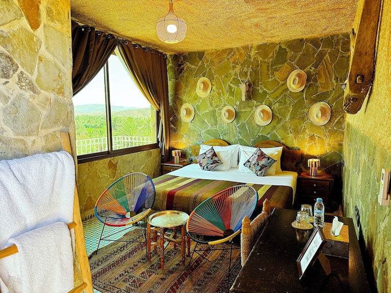 Riad Chay Boutique bedroom with rustic stone wall, double bed with colorful striped blanket, two multicolor chairs, and large window