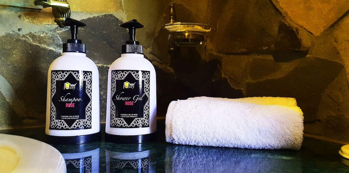 Riad Chay Boutique branded white shampoo and rose shower gel bottles with black pump tops alongside a rolled white towel on a stone countertop