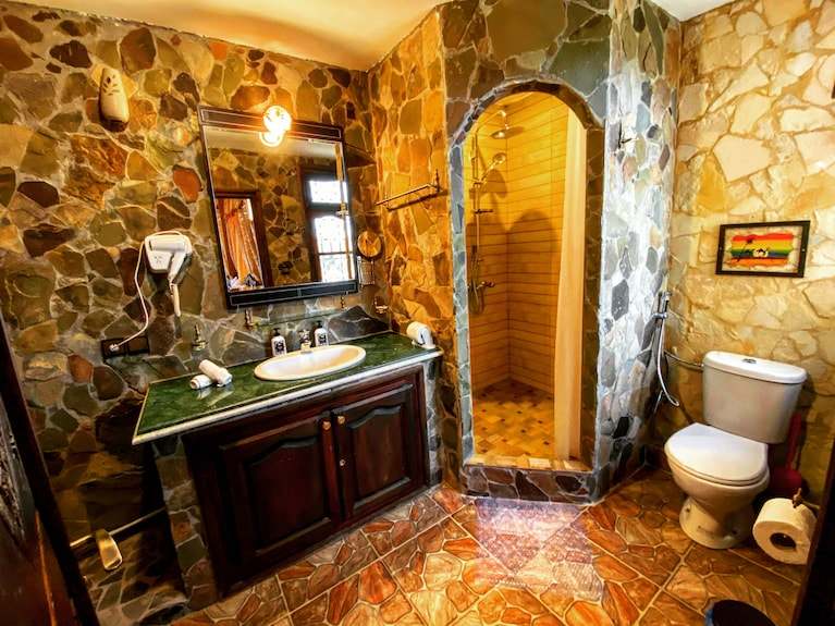 Bathroom with stone-textured walls, tiled floor, wooden vanity with green countertop, and arched shower space in Riad Chay Boutique