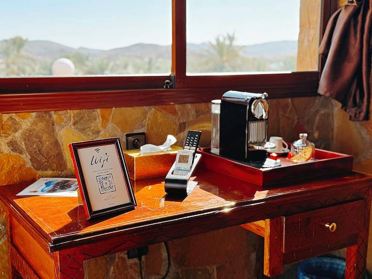 Riad Chay Boutique wooden desk with stone wall backdrop, coffee machine on tray, phone, tissue box, and framed WiFi sign
