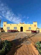 Riad Dar Menorca yellow stucco building with red accents and tiled outdoor seating under a blue sky in Merzouga