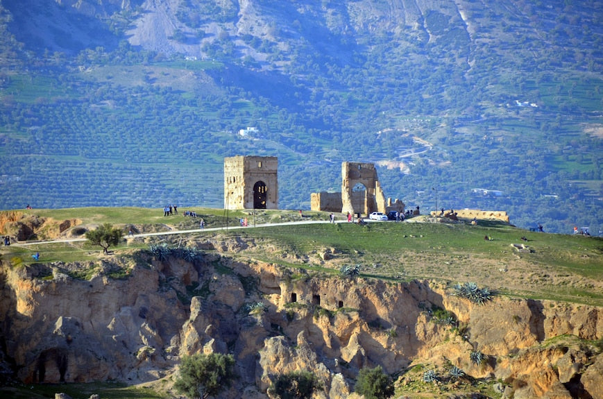 Ancient stone ruins on a grassy hilltop with a mountainous backdrop under a clear sky, showcasing historic architecture
