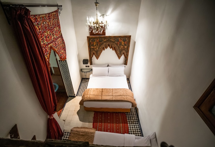 Riad Dar Tahri SPA bedroom with white walls, double bed, decorative wooden headboard, patterned red wall hanging, and a chandelier
