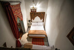 Riad Dar Tahri SPA bedroom with white walls, double bed, decorative wooden headboard, patterned red wall hanging, and a chandelier