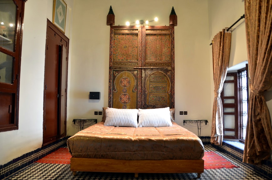 Riad Dar Tahri SPA bedroom with traditional Moroccan carved wooden headboard, patterned floor tiles, and brown velvet curtains