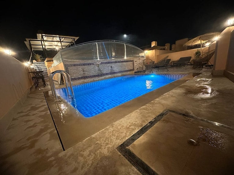 Outdoor rectangular blue-lit swimming pool at night with stone deck and lounge chairs under canopy at Riad Dar Tahri SPA