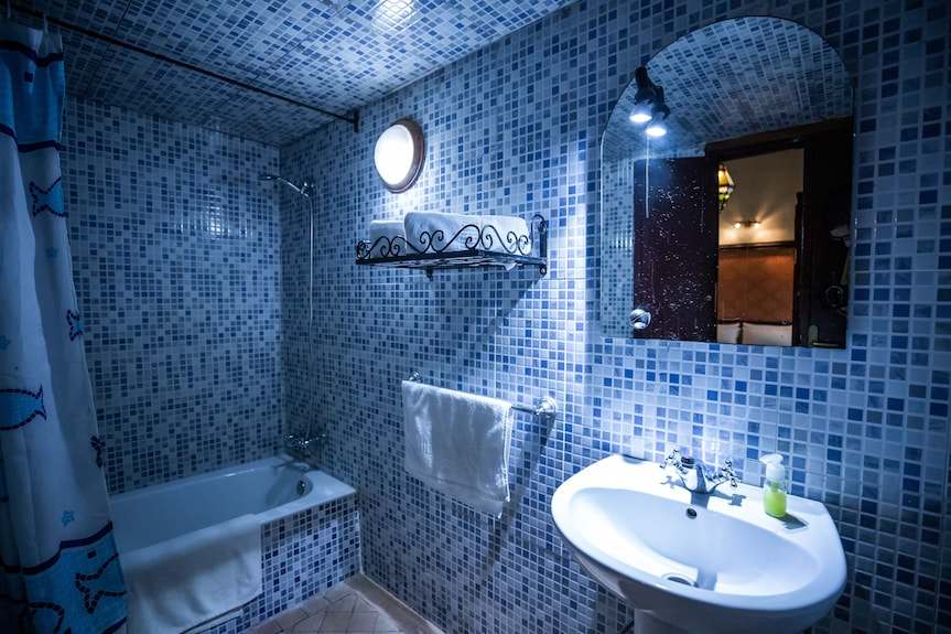 Riad Dar Tahri SPA bathroom with blue mosaic tiled walls, white porcelain sink, bathtub, towel rack, and round wall light