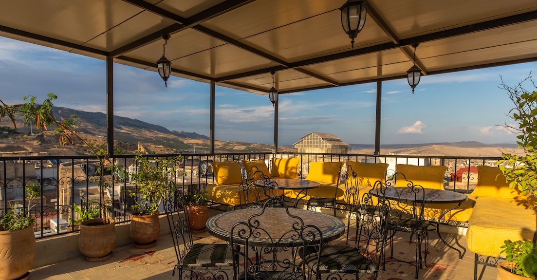 Riad Dar Tahri SPA covered terrace with black wrought iron tables and chairs and yellow cushioned benches overlooking scenic hills