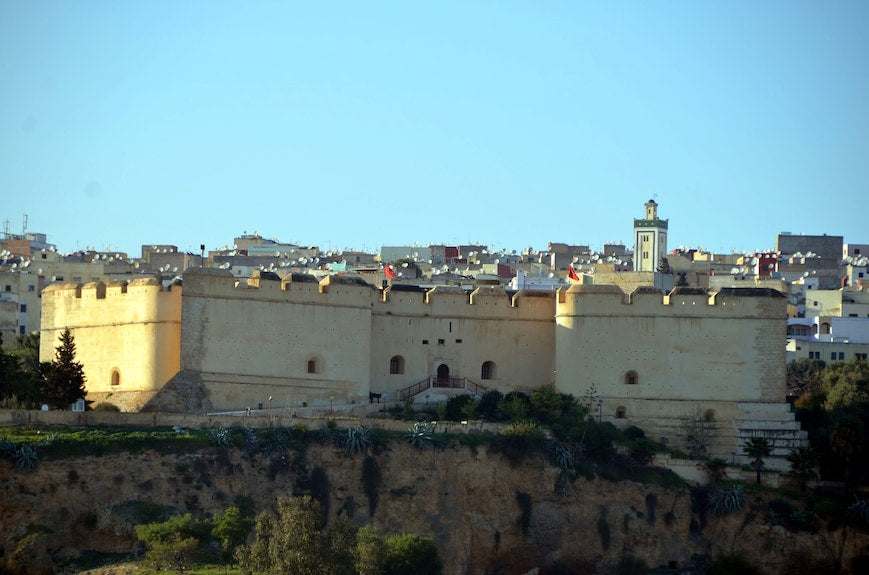Historic beige stone fortress with crenellated walls situated on a cliff with city buildings behind under clear blue sky, representing Riad Dar Tahri SPA surroundings