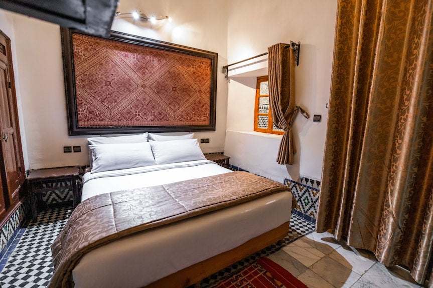 Riad Dar Tahri SPA Moroccan bedroom with white bedding, patterned wooden headboard, brown decorative throw and curtains, traditional tile floor