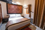 Riad Dar Tahri SPA Moroccan bedroom with white bedding, patterned wooden headboard, brown decorative throw and curtains, traditional tile floor
