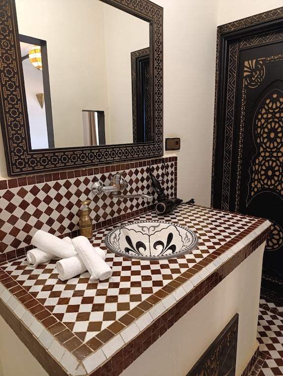 Riad La Luna decorative bathroom sink with brown and white tiled countertop, ornate mirror frame, and black patterned door