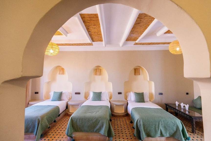 Riad La Luna bedroom with three single beds featuring light green throws, white linens, beige walls, and arched recesses