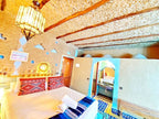 Riad La Luna bedroom in Merzouga Morocco with traditional decor, beige textured walls, wooden beams, patterned pillows, and tiled bathroom