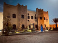 Riad La Luna with beige and stone textured walls, traditional Moroccan design, and illuminated exterior at dusk with palm trees nearby.