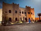Riad La Luna with beige and stone textured walls, traditional Moroccan design, and illuminated exterior at dusk with palm trees nearby.