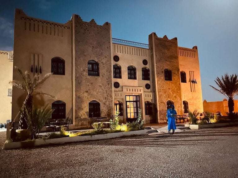 Riad La Luna with beige and stone textured walls, traditional Moroccan design, and illuminated exterior at dusk with palm trees nearby.