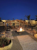 Riad La Luna outdoor seating area at night with fire pit, wooden pergola, cushioned benches, and gravel flooring under clear blue sky