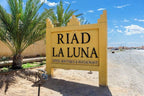 Riad La Luna large yellow stone sign with black text at desert entrance and palm trees nearby under blue sky
