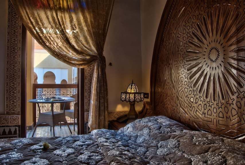 Riad Salam Fes bedroom with ornate carved wooden headboard, patterned blue comforter, decorative lamp, and balcony with seating
